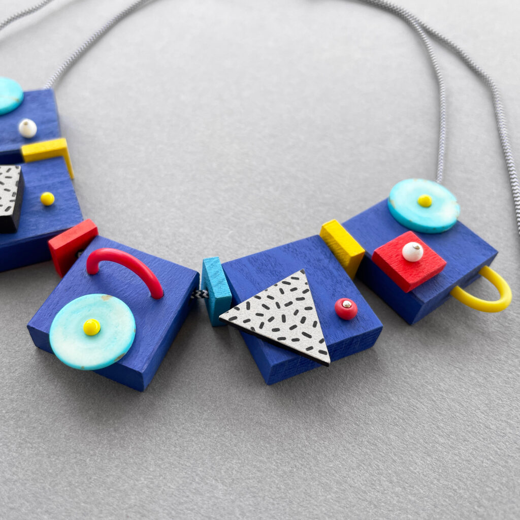 Blue Memphis milano abstract wooden statement necklace with turquoise, red, yellow and black-and-white details on a grey background